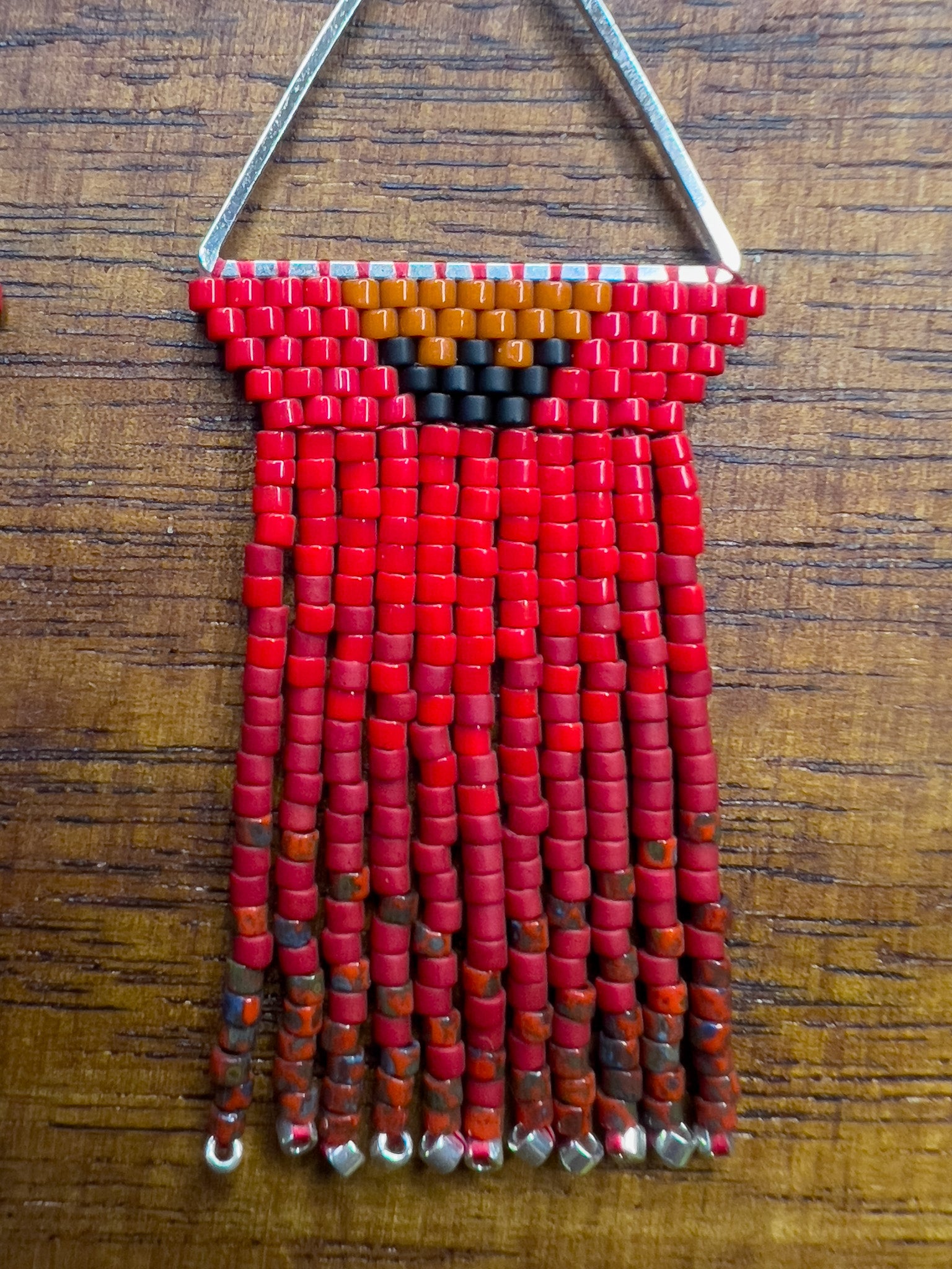 Close up of Northern Cardinal inspired earrings on a wooden background, with a triangular silver frame holding rows of red beads and an orange and black accent at the top, leading to long red fringe that ends with silver bead accents.