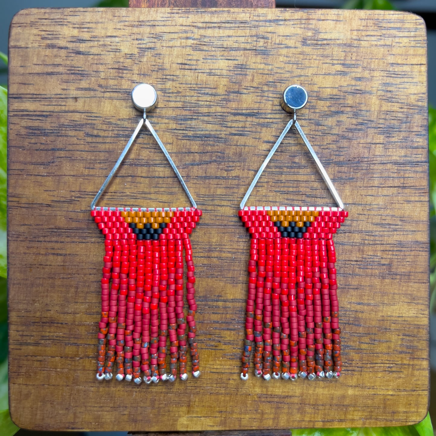 Northern Cardinal inspired earrings on a wooden background, with a triangular silver frame holding rows of red beads and an orange and black accent at the top, leading to long red fringe that ends with silver bead accents.