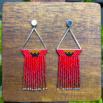 Northern Cardinal inspired earrings on a wooden background, with a triangular silver frame holding rows of red beads and an orange and black accent at the top, leading to long red fringe that ends with silver bead accents.