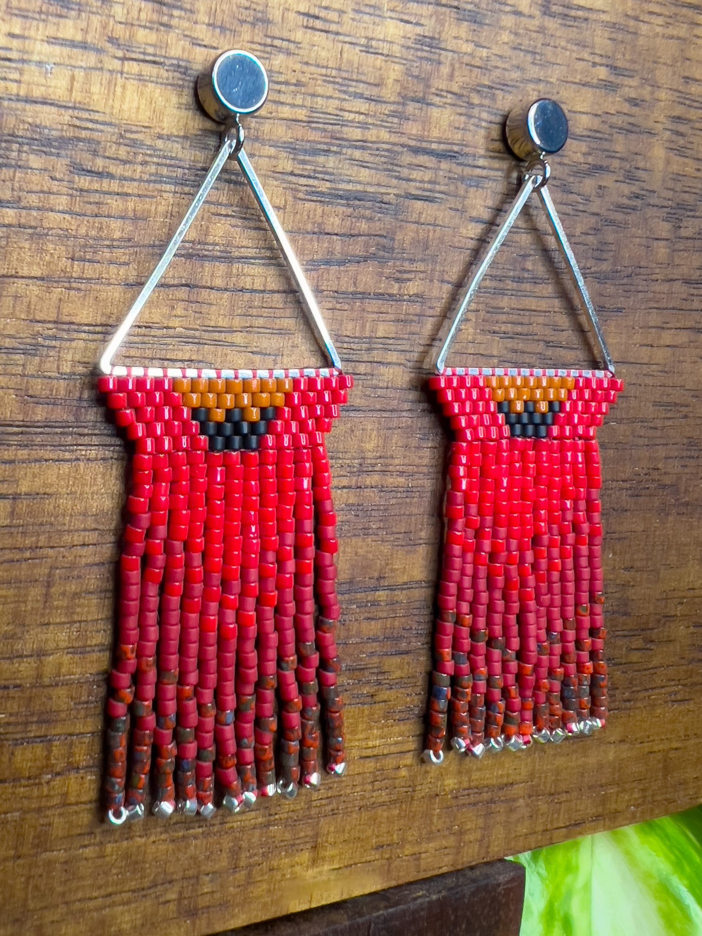 Side view of Northern Cardinal inspired earrings on a wooden background, with a triangular silver frame holding rows of red beads and an orange and black accent at the top, leading to long red fringe that ends with silver bead accents.