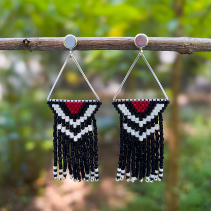 Pair of beaded Pileated Woodpecker earrings hanging from a branch, with a triangular silver frame holding a red, white, and black chevron pattern at the top, followed by cascading shiny and matte black fringe ending with silver beads.