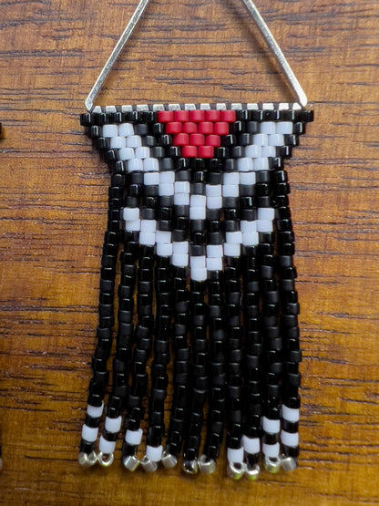 Close up of Pileated Woodpecker inspired earrings on a wooden background, featuring a silver triangular frame with a red, white, and black chevron design at the top, leading into shiny and matte black fringe with silver bead accents.