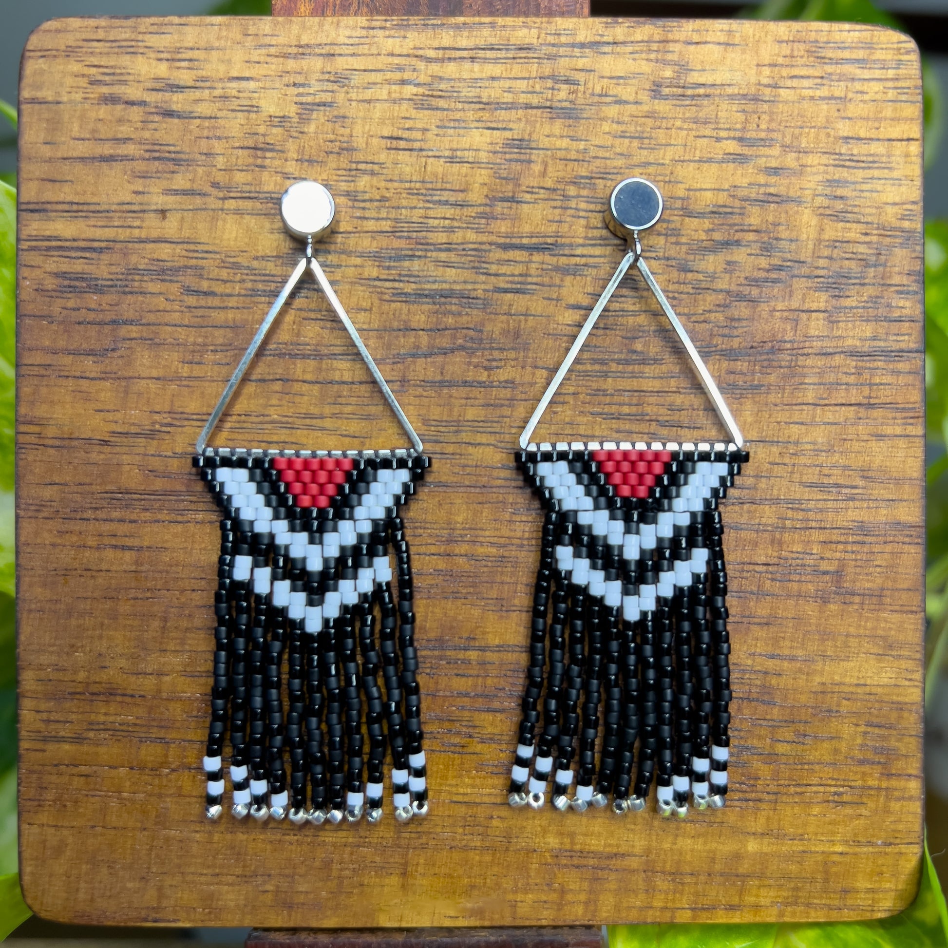 Pileated Woodpecker inspired earrings on a wooden background, featuring a silver triangular frame with a red, white, and black chevron design at the top, leading into shiny and matte black fringe with silver bead accents.