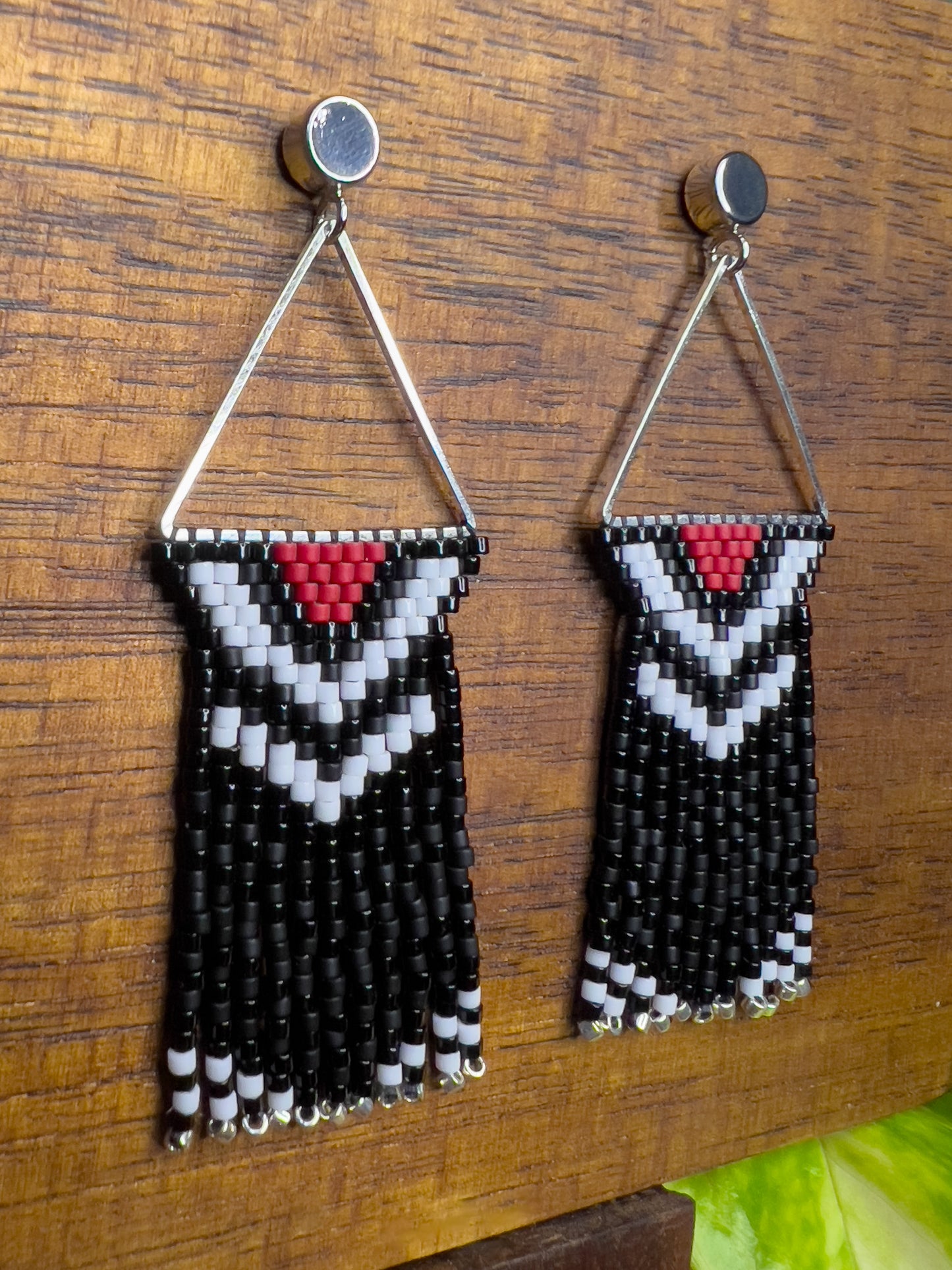 Side view of Pileated Woodpecker inspired earrings on a wooden background, featuring a silver triangular frame with a red, white, and black chevron design at the top, leading into shiny and matte black fringe with silver bead accents.