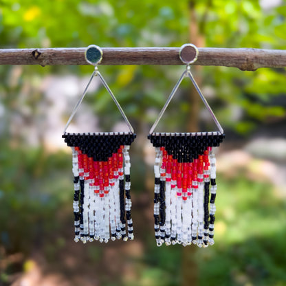 Pair of beaded earrings hanging from a branch, featuring a triangular silver frame with black, red, and white beading that resembles the markings of a Rose Breasted Grosbeak, with fringe in ending in silver beads.