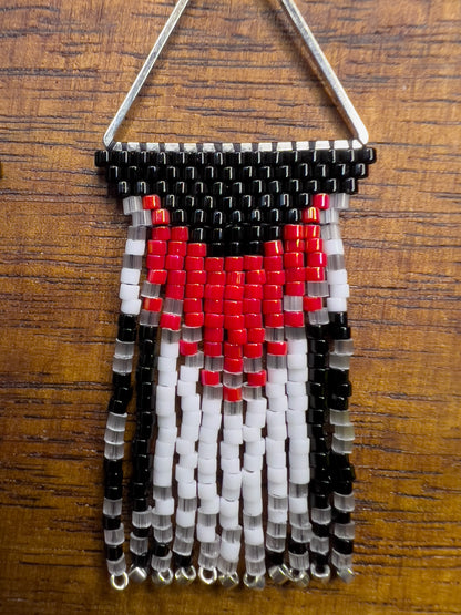 Close up of Rose Breasted Grosbeak inspired earrings on a wooden background, showcasing a silver triangular frame holding a bold red and white design against a black base, leading into a layered fringe of black, white, and clear beads.