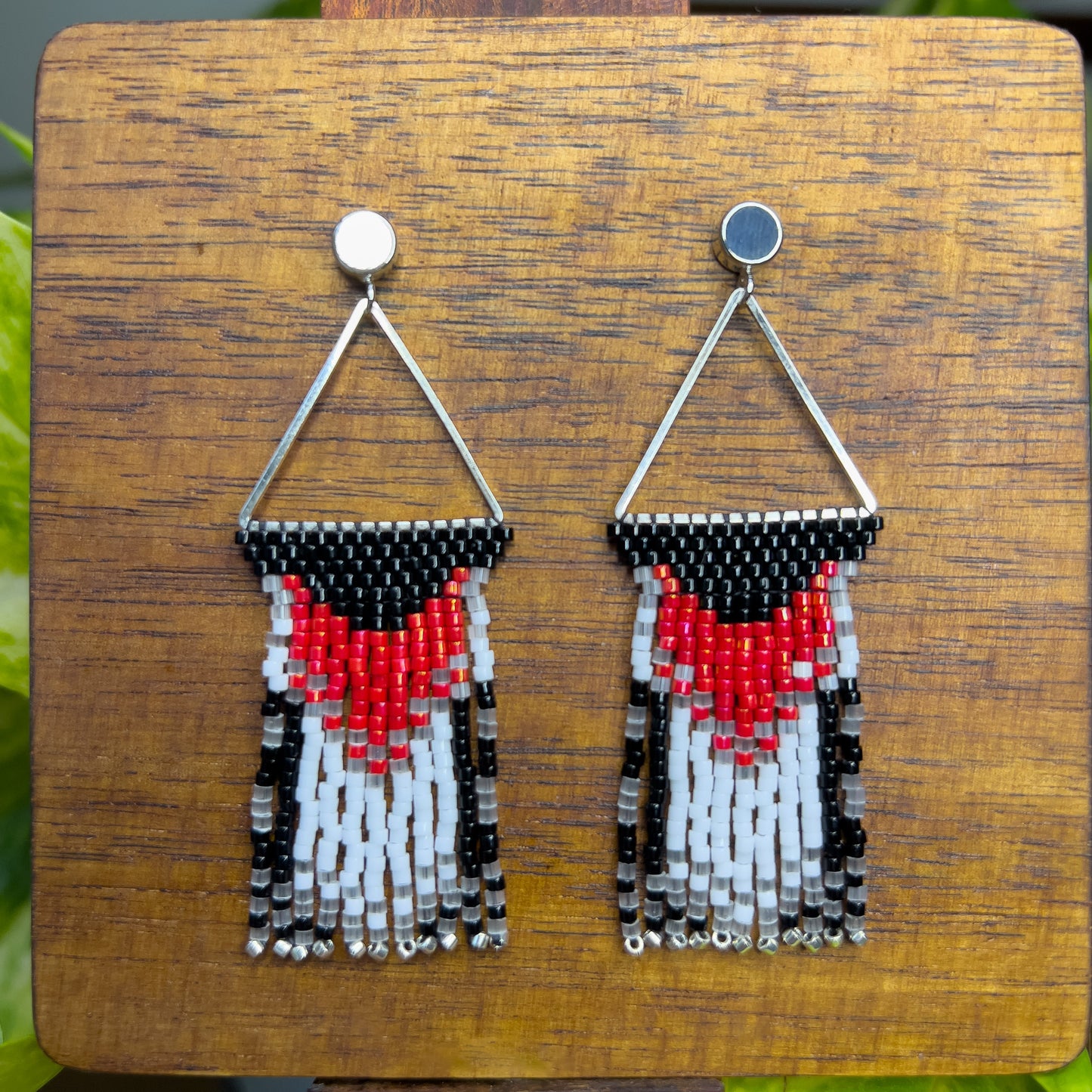 Rose Breasted Grosbeak inspired earrings on a wooden background, showcasing a silver triangular frame holding a bold red and white design against a black base, leading into a layered fringe of black, white, and clear beads.