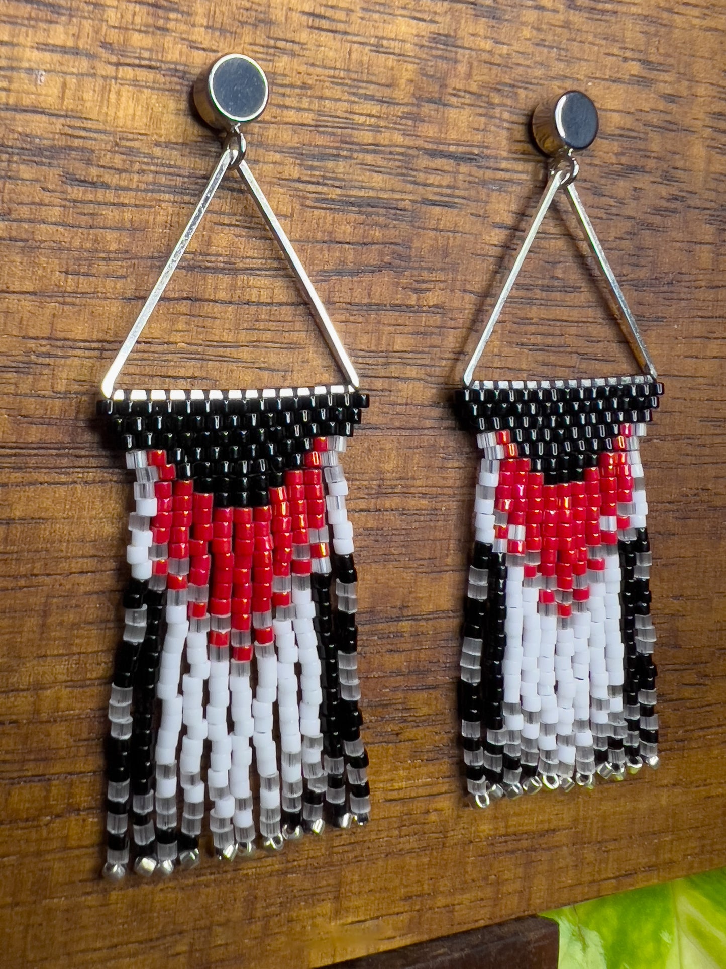 Side view of Rose Breasted Grosbeak inspired earrings on a wooden background, showcasing a silver triangular frame holding a bold red and white design against a black base, leading into a layered fringe of black, white, and clear beads.