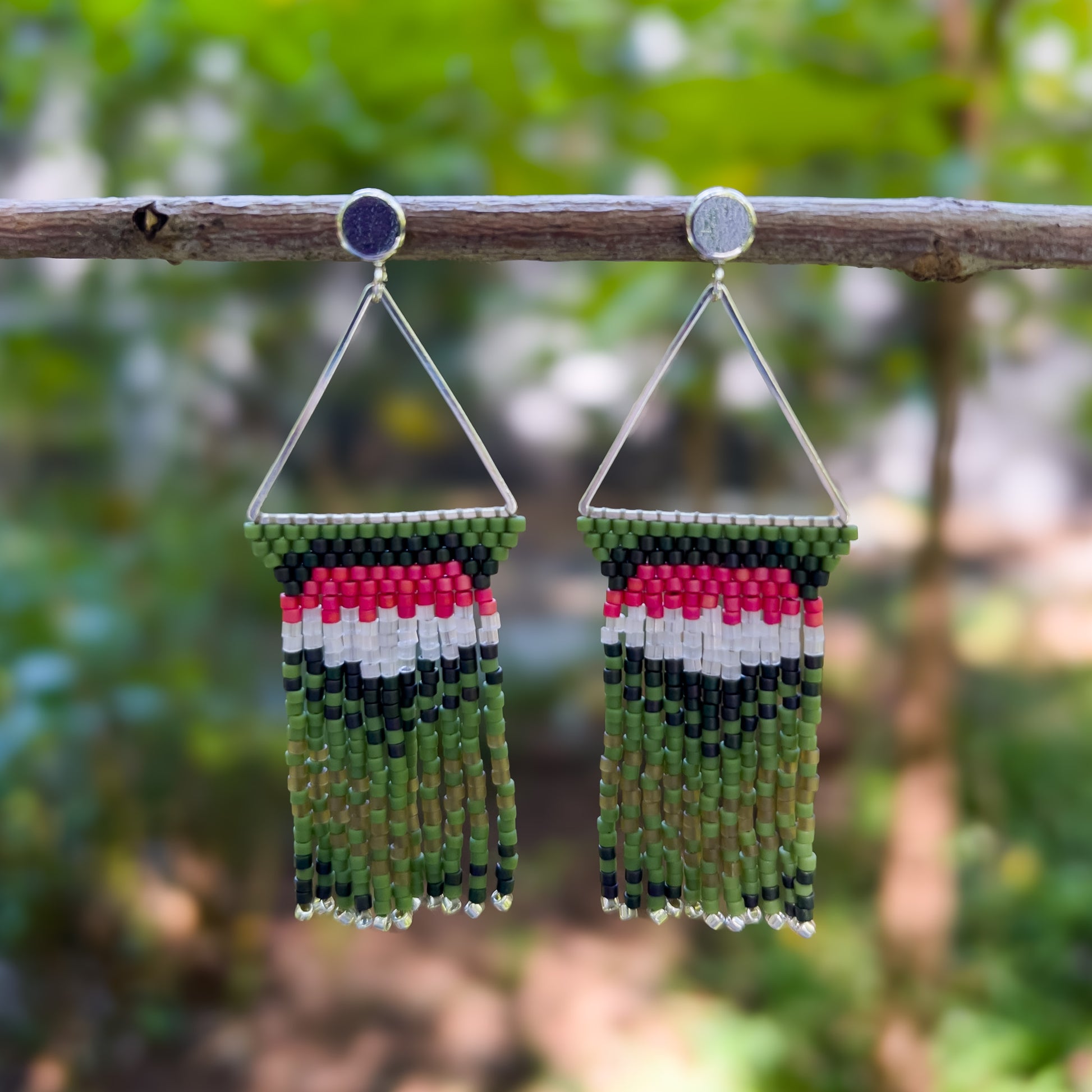 Pair of beaded earrings hanging from a branch, featuring a triangular silver frame with green, pink, white, and dark green beading that mimics the colors of a Ruby Throated Hummingbird, cascading into green fringe with silver ends.