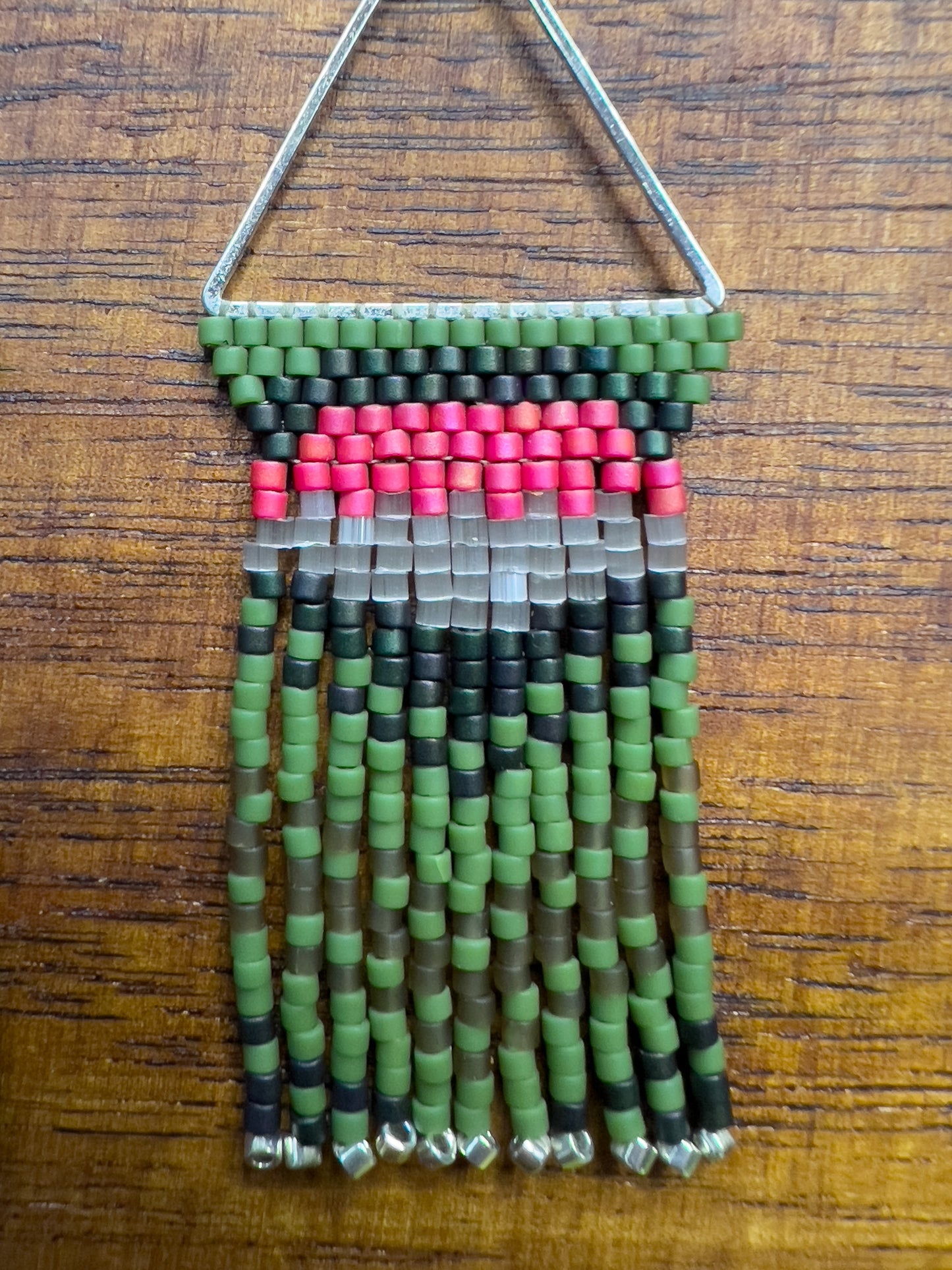 Close up of Ruby Throated Hummingbird inspired earrings on a wooden background, showcasing a triangular frame with rows of green, pink, and gray beads at the top, leading into long green fringe accented with silver ends.