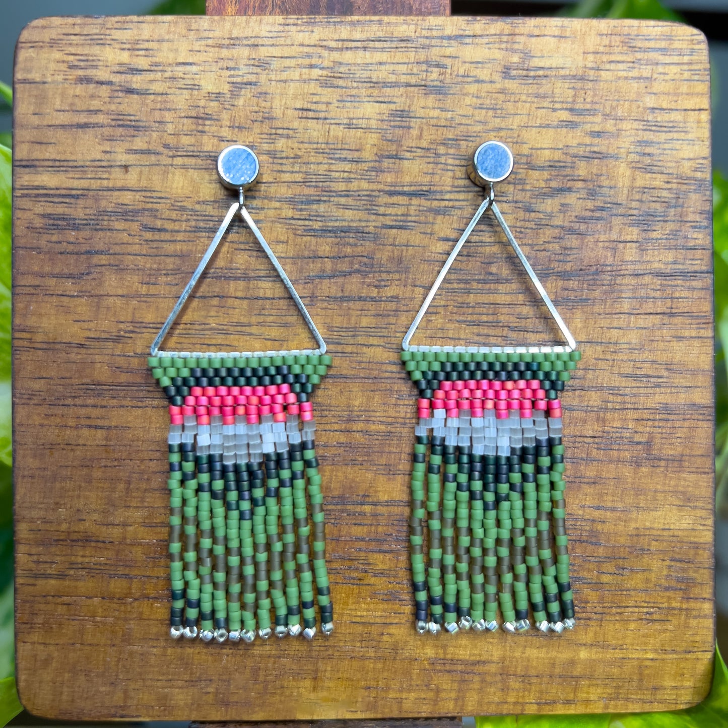 Ruby Throated Hummingbird inspired earrings on a wooden background, showcasing a triangular frame with rows of green, pink, and gray beads at the top, leading into long green fringe accented with silver ends.