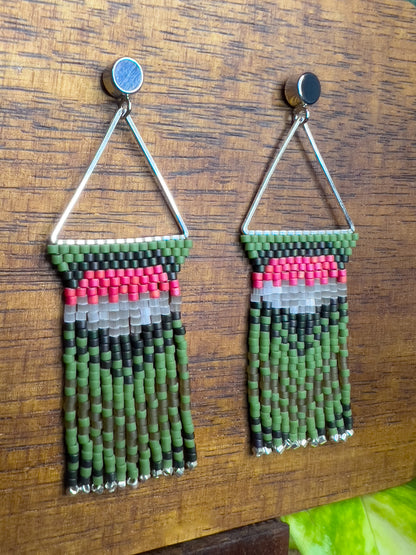 Side view of Ruby Throated Hummingbird inspired earrings on a wooden background, showcasing a triangular frame with rows of green, pink, and gray beads at the top, leading into long green fringe accented with silver ends.