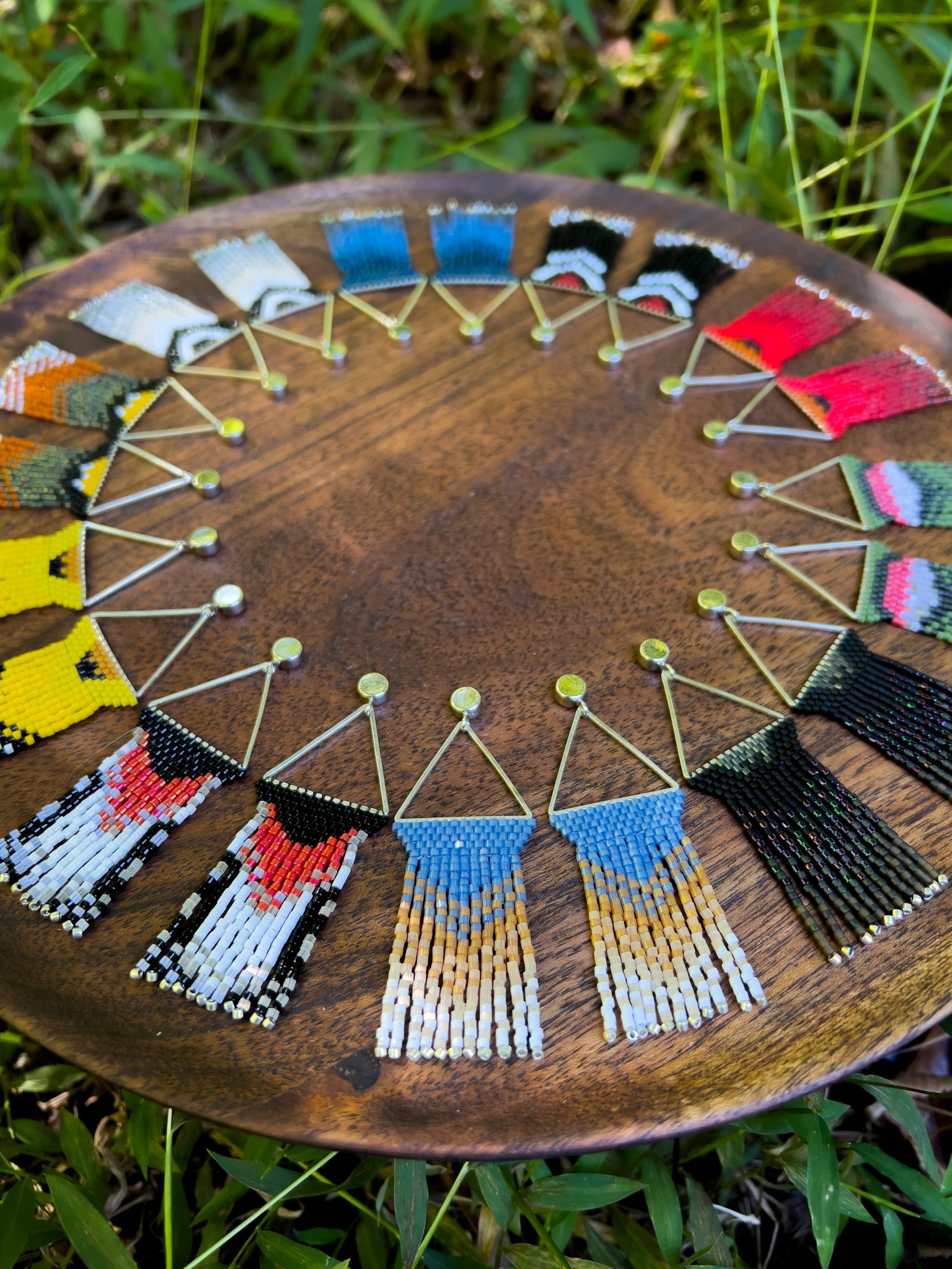 Handcrafted bird-inspired beaded earrings arranged in a semi-circle on a wooden tray, with intricate beadwork in various colors, against a backdrop of lush greenery.