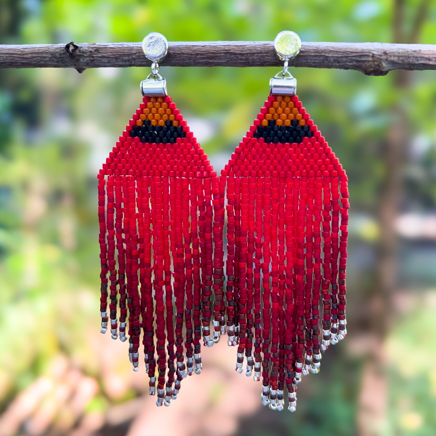 Pair of vibrant red Northern Cardinal beaded earrings hanging from a branch, featuring a triangular design with an orange and black accent at the top, resembling a cardinal's head, with long red fringe tapering into silver bead accents.