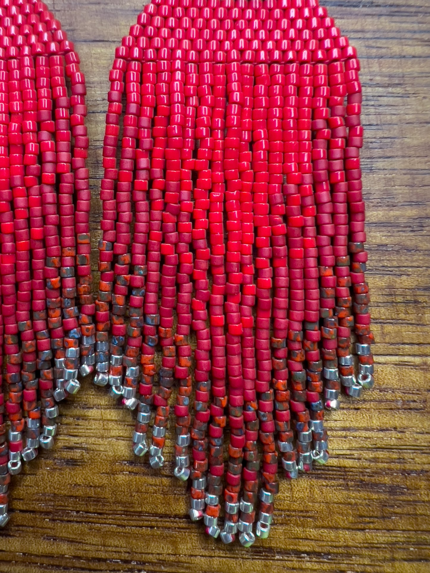 Close up of vibrant red beaded Northern Cardinal earrings on a wooden background, showcasing an orange and black beaded section at the top, followed by cascading red fringe that fades into subtle silver bead accents at the bottom.