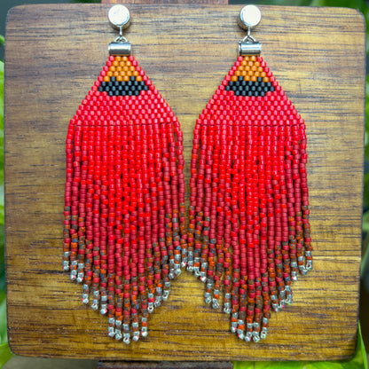 Vibrant red beaded Northern Cardinal earrings on a wooden background, showcasing an orange and black beaded section at the top, followed by cascading red fringe that fades into subtle silver bead accents at the bottom.