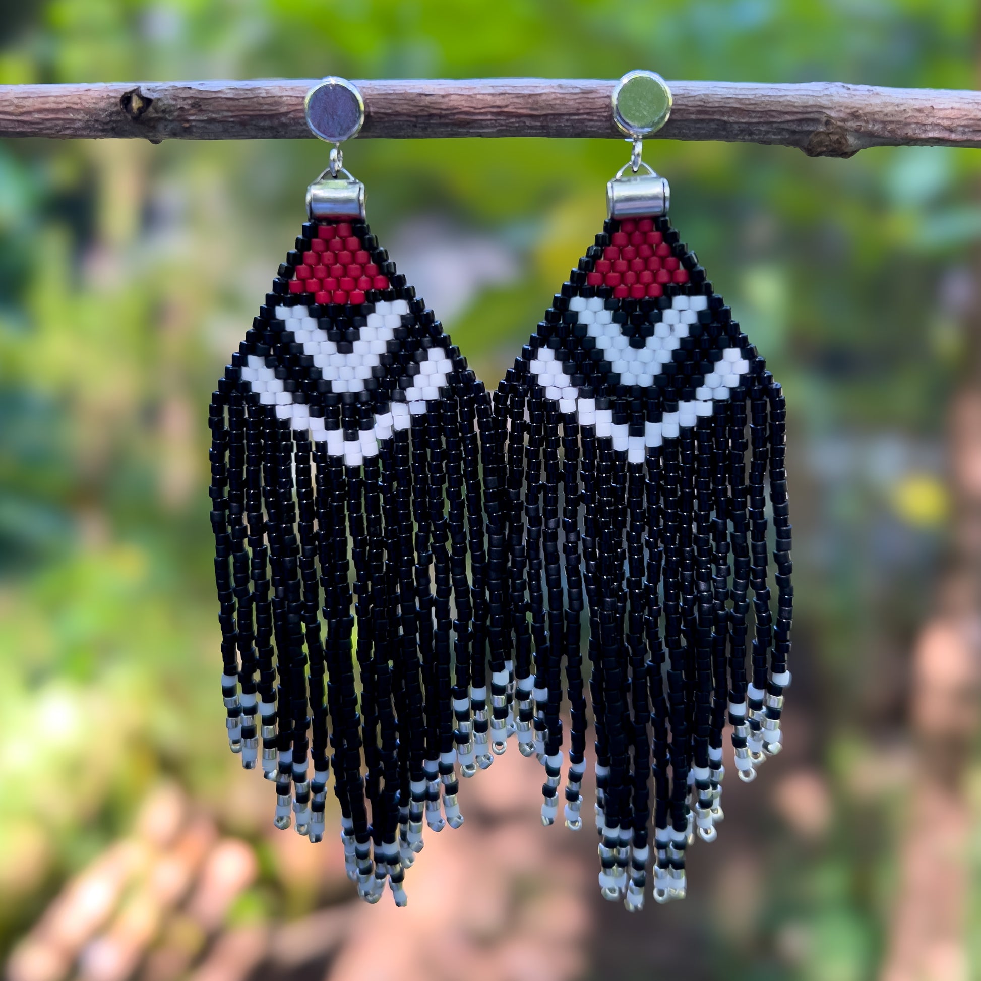 Pair of beaded Pileated Woodpecker earrings hanging from a branch, with a red, white, and black geometric design at the top that transitions into long shiny and matte black fringe accented by silver beads at the ends.