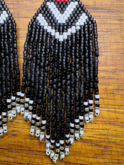 Close up of beaded Pileated Woodpecker earrings on a wooden background, showcasing a red triangular section with white chevron patterns on black beading, leading to shiny and matte black fringe ending in silver bead accents.