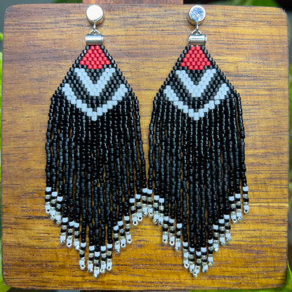 Beaded Pileated Woodpecker earrings on a wooden background, showcasing a red triangular section with white chevron patterns on black beading, leading to shiny and matte black fringe ending in silver bead accents.