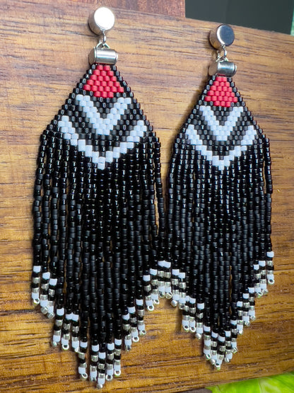 Side view of beaded Pileated Woodpecker earrings on a wooden background, showcasing a red triangular section with white chevron patterns on black beading, leading to shiny and matte black fringe ending in silver bead accents.