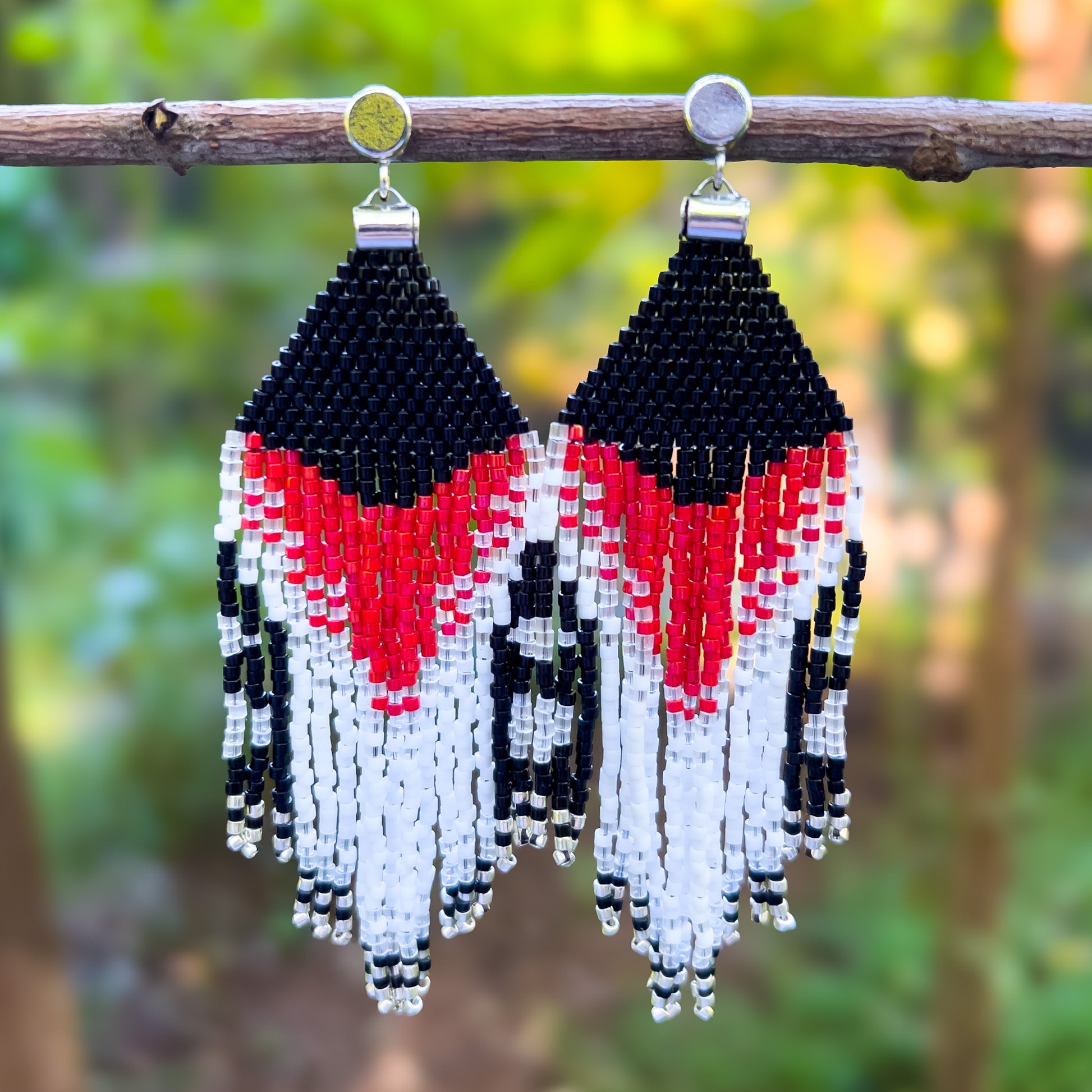 Pair of beaded earrings hanging from a branch, inspired by the Rose Breasted Grosbeak, featuring a black beaded top with a bold red and white chevron design, flowing into shimmering fringe with black, red, white, and clear beads.