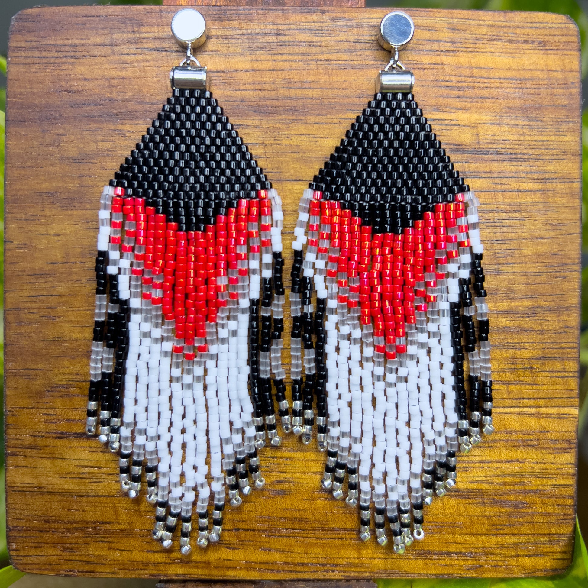 Rose Breasted Grosbeak inspired earrings on a wooden background, showing a striking red and white chevron pattern against a black beaded top, with long, cascading fringe in black, white, and clear beads.