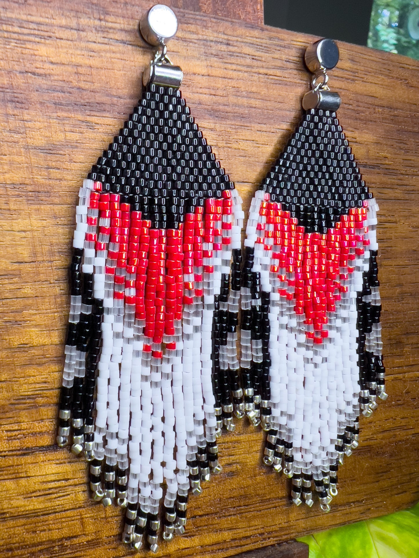 Side view of Rose Breasted Grosbeak inspired earrings on a wooden background, showing a striking red and white chevron pattern against a black beaded top, with long, cascading fringe in black, white, and clear beads.