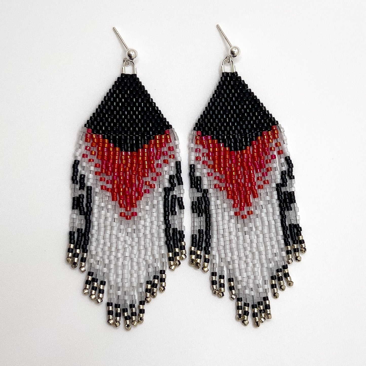 Pair of beaded earrings displayed on a white background, inspired by the Rose Breasted Grosbeak, featuring a black beaded top with a bold rose and white chevron design, flowing into shimmering fringe with black, red, white, and clear beads.