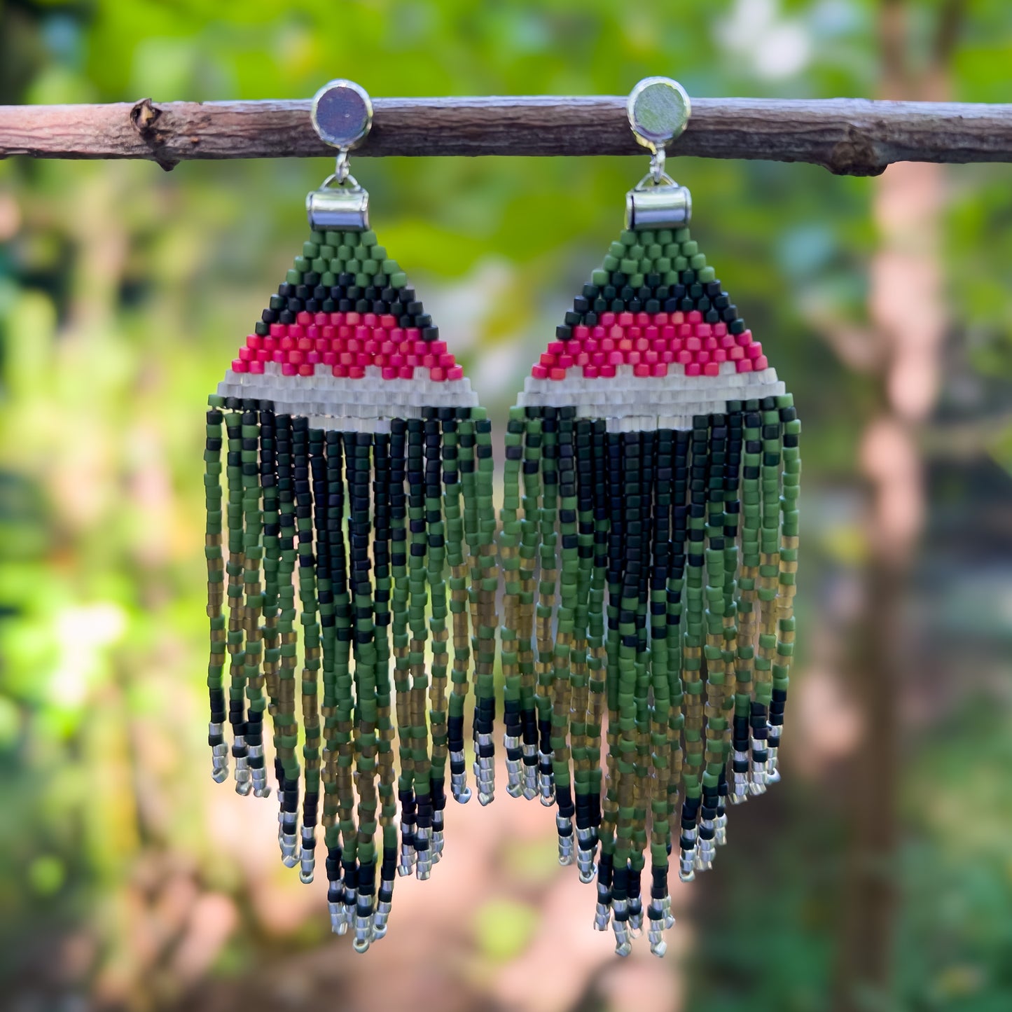 Pair of beaded earrings hanging from a branch, inspired by the Ruby Throated Hummingbird, featuring a bold pink, white, and green beaded design at the top that flows into layered green and brown fringe with silver bead tips.