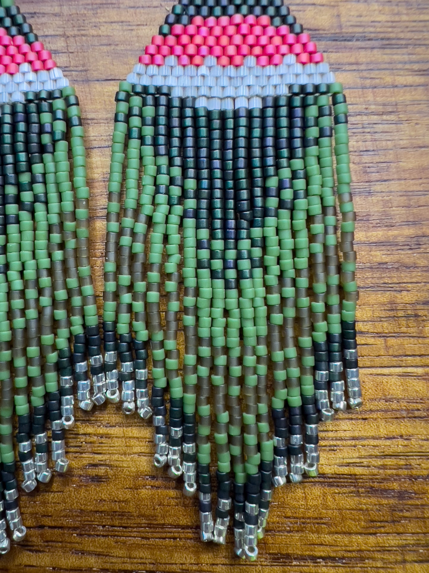 Close up of Ruby Throated Hummingbird inspired earrings on a wooden background, showcasing a vibrant pink and white stripe above dark green beading, with cascading green fringe accented by silver beads at the ends.