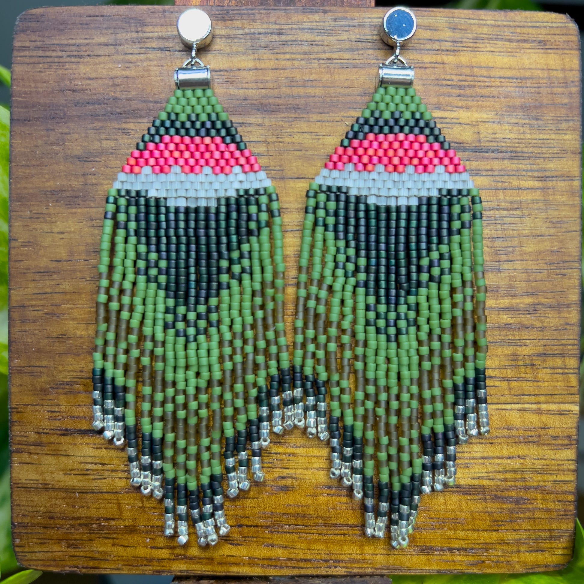 Ruby Throated Hummingbird inspired earrings on a wooden background, showcasing a vibrant pink and white stripe above dark green beading, with cascading green fringe accented by silver beads at the ends.