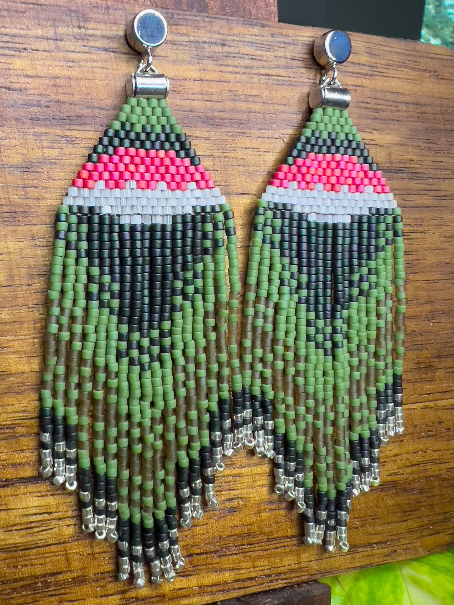 Side view of Ruby Throated Hummingbird inspired earrings on a wooden background, showcasing a vibrant pink and white stripe above dark green beading, with cascading green fringe accented by silver beads at the ends.